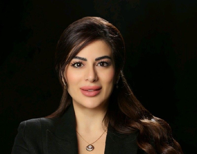 The image is a professional headshot of Ghenwa Habbal. She is standing against a neutral background, and her expression is confident and friendly. She is wearing business attire and is holding a book titled "Shaken Stirred But Not Deterred." The book's cover features vibrant colors and bold typography. Ghenwa's posture exudes a sense of empowerment and determination.